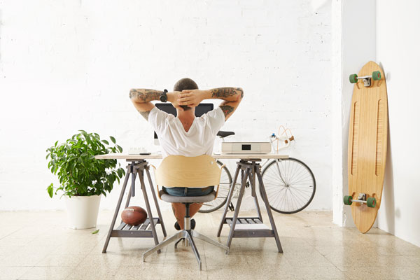 A freelance graphic designer sits relaxed at a minimalist workspace, hands behind their head, embracing the freedom of remote work. The creative workspace features a modern desk, a bicycle, a skateboard, and a potted plant, symbolizing the flexibility and personal touch that comes with freelancing.